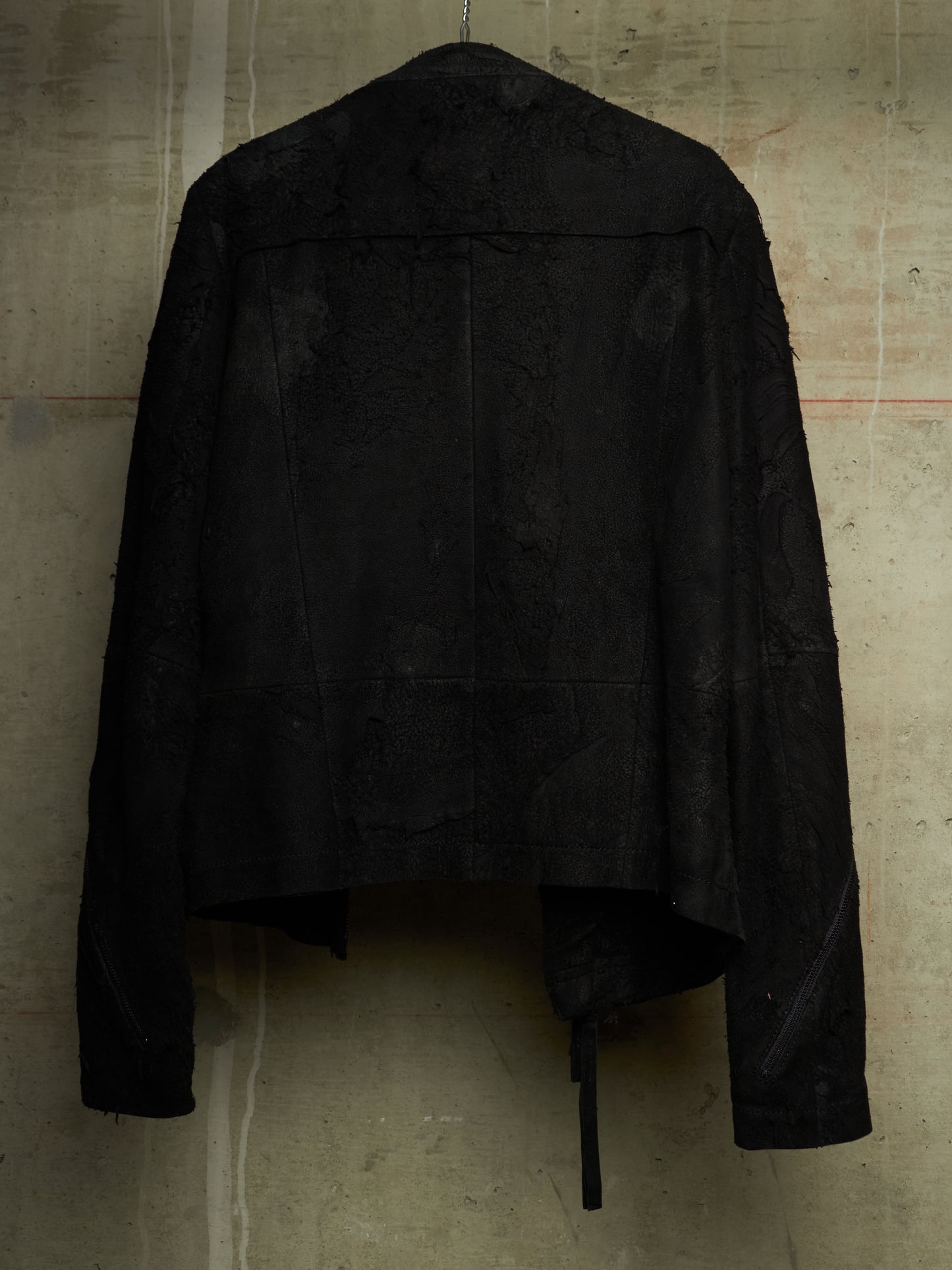 A black textured jacket with a heavily distressed, frayed, and tattered design, hanging against a concrete wall.