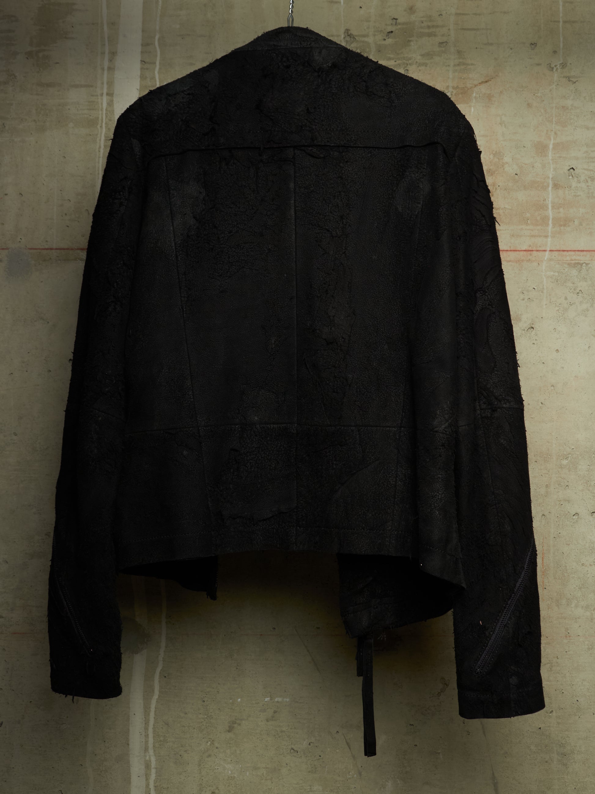 A black textured jacket with a heavily distressed, frayed, and tattered design, hanging against a concrete wall.