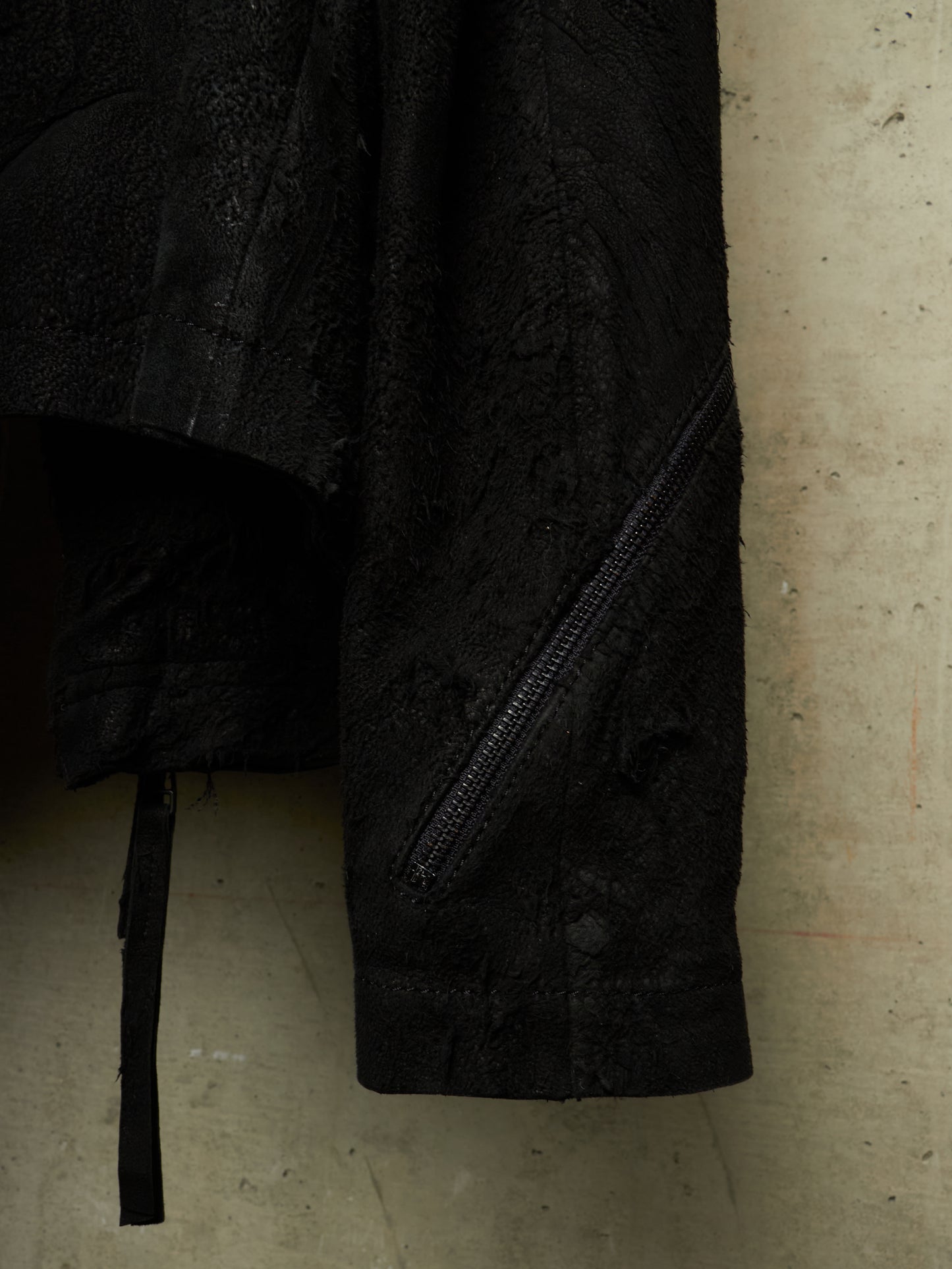 Close-up of a black textured jacket or coat with a diagonal zip pocket and a hanging black strap, photographed against a concrete wall.