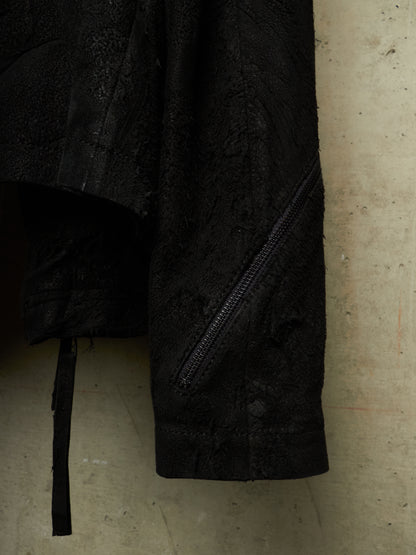 Close-up of a black textured jacket or coat with a diagonal zip pocket and a hanging black strap, photographed against a concrete wall.