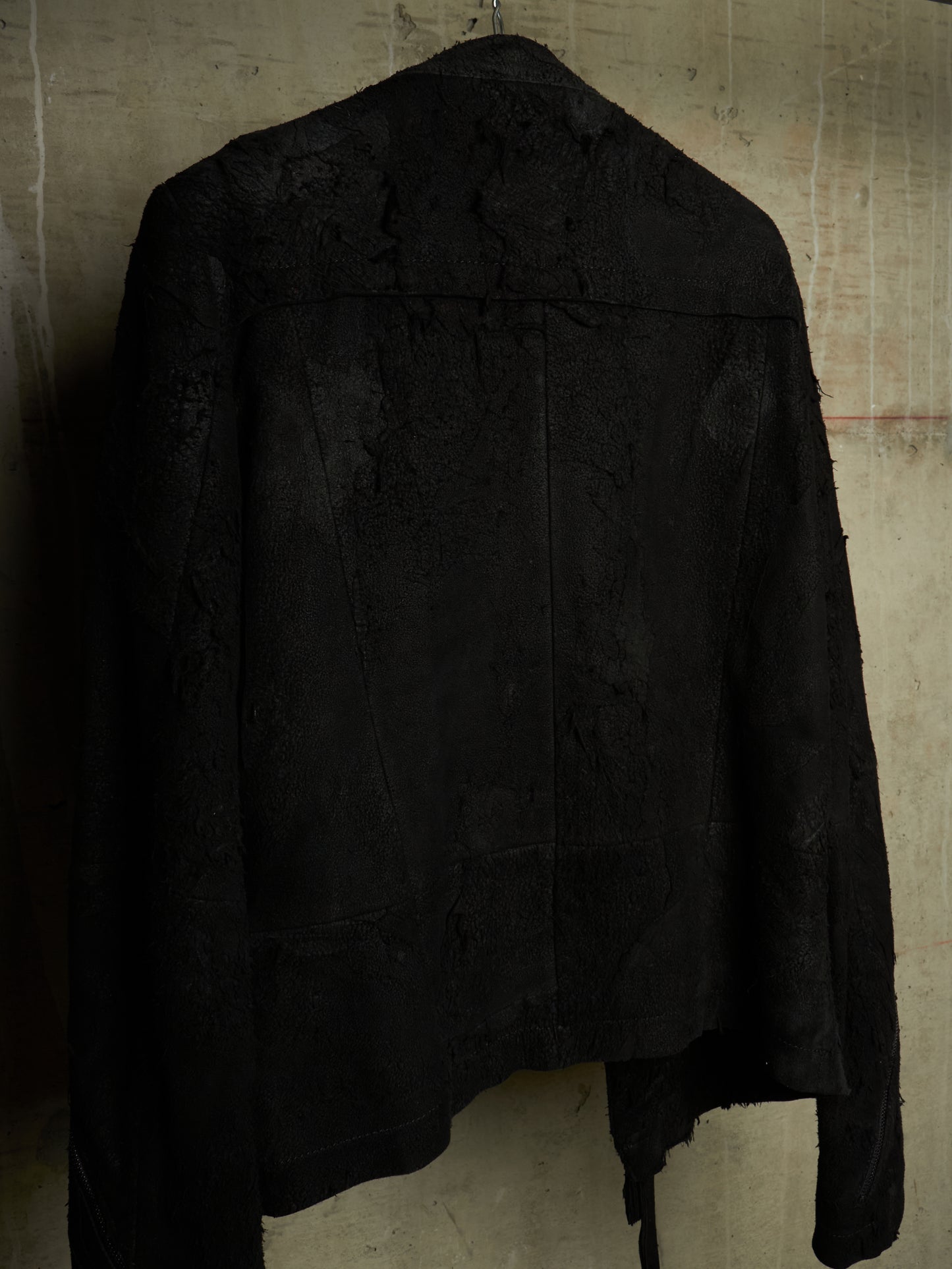 A black textured jacket with a distressed, tattered fabric finish, hanging against a textured concrete wall.