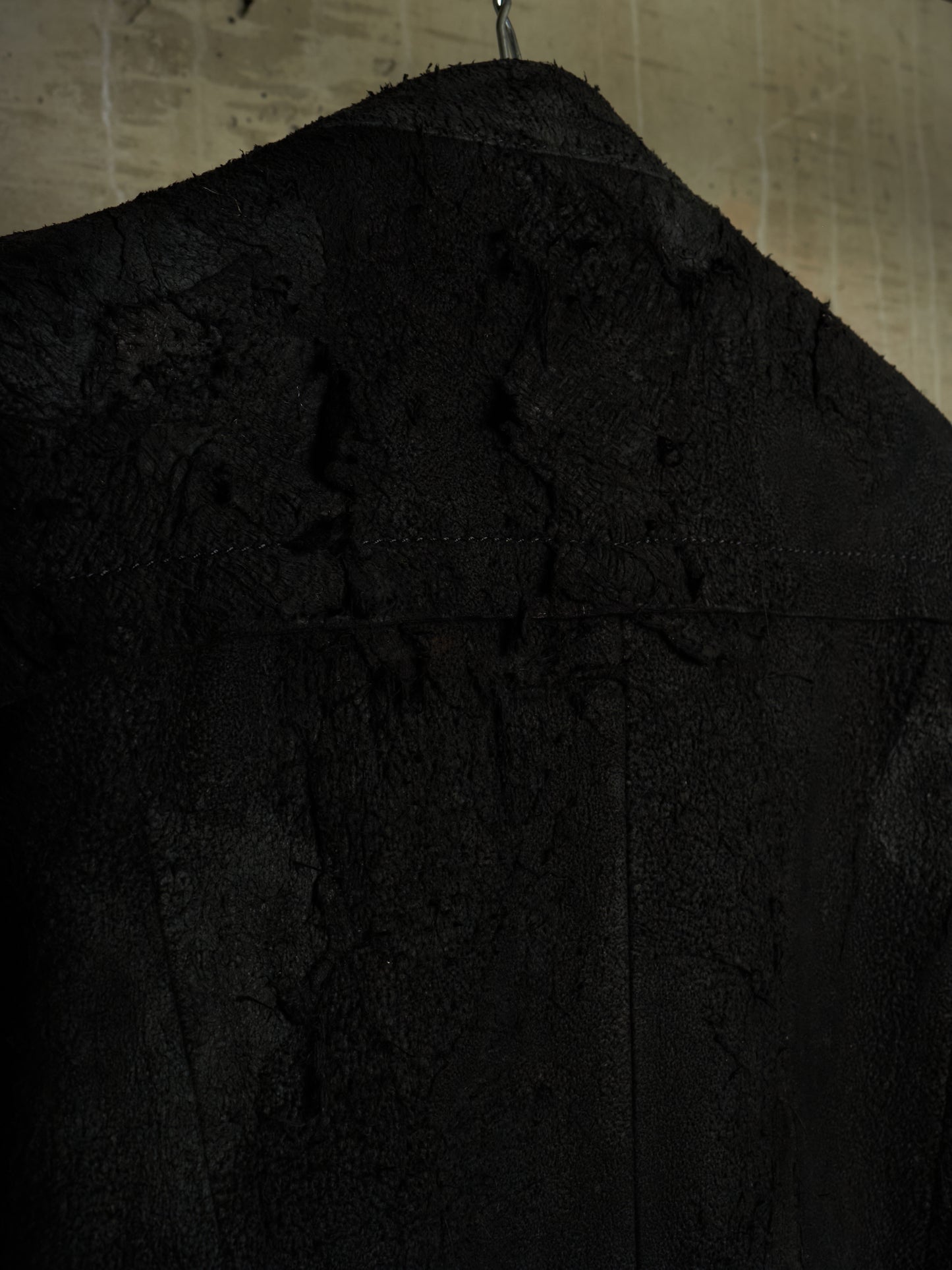 Close-up of a black textured jacket or coat hanging against a dimly lit beige wall, showing a heavily distressed, frayed, and raw fabric surface.