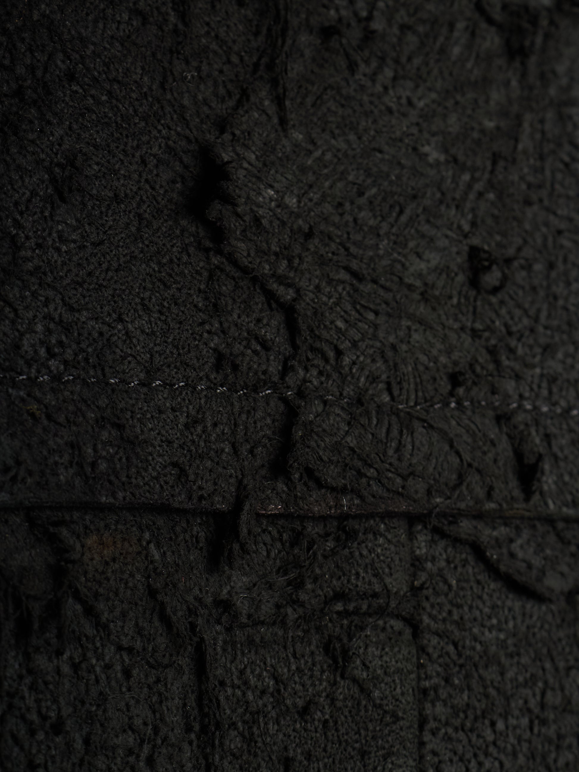 A close-up of a dark textured material with a heavily distressed, cracked, and weathered appearance, showing fine grain and deep shadows.