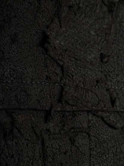 A close-up of a dark textured material with a heavily distressed, cracked, and weathered appearance, showing fine grain and deep shadows.