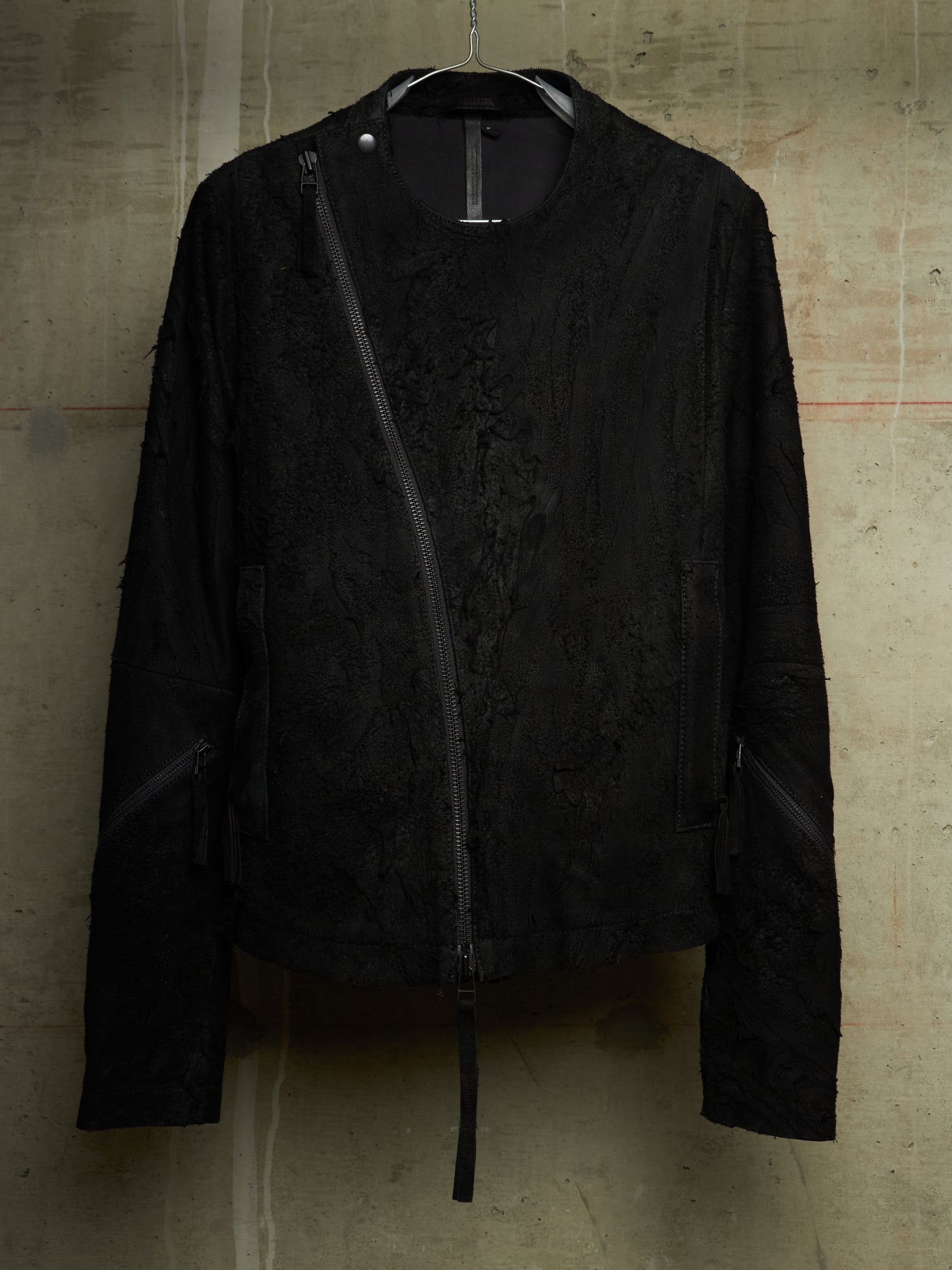 A black textured jacket with an asymmetrical zipper, hanging on a black hanger against a concrete wall.