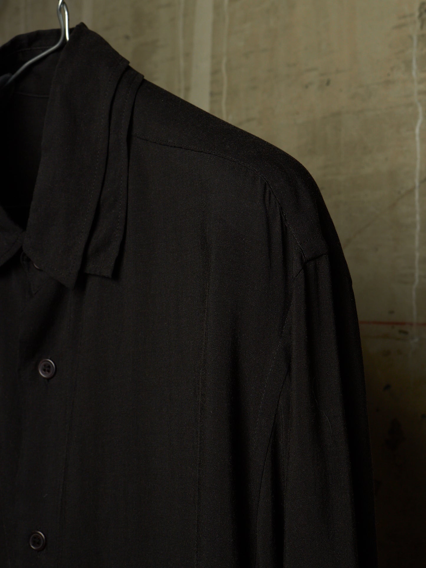 A close-up of a black long-sleeve button-up shirt hanging on a metal hanger against a textured concrete wall.