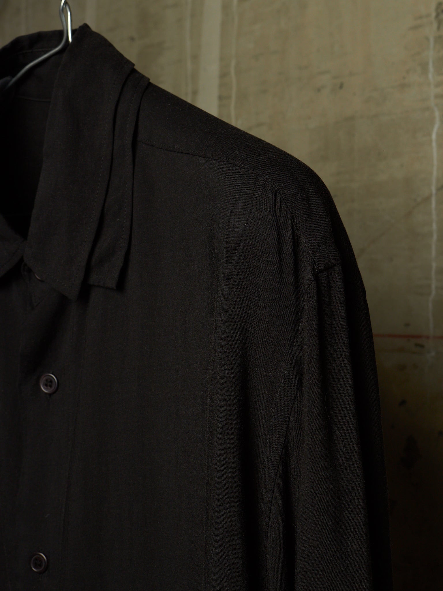 A close-up of a black long-sleeve button-up shirt hanging on a metal hanger against a textured concrete wall.