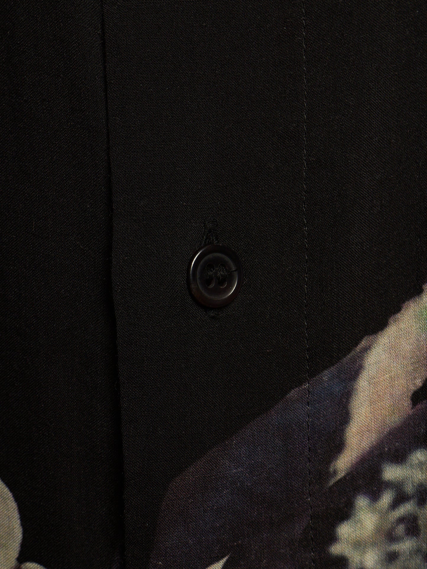 Close-up detail of a black button-up shirt featuring a single dark button and a subtle abstract floral pattern visible in the lower right corner.
