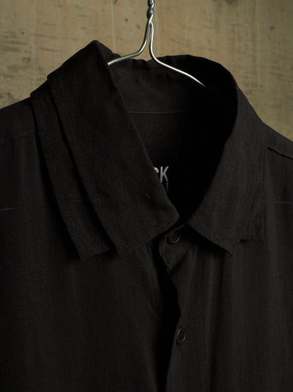 A close-up of a black button-front shirt hanging on a metal hanger against a textured beige wall.