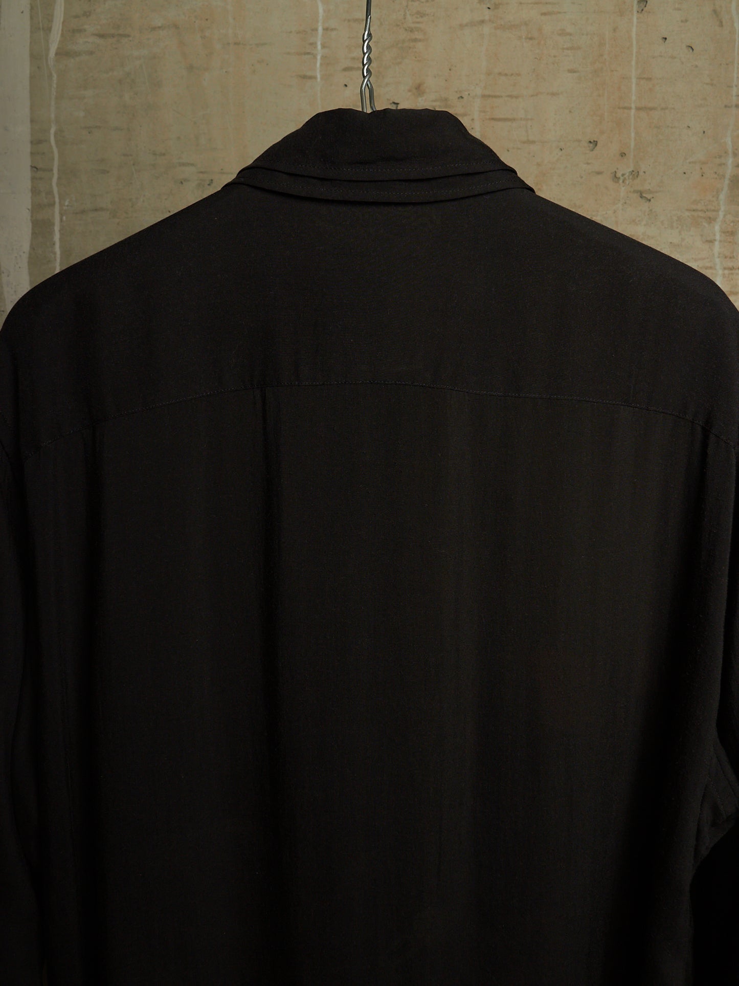 A black long-sleeve collared shirt hanging on a metal hanger against a textured beige wall, shown from the back.