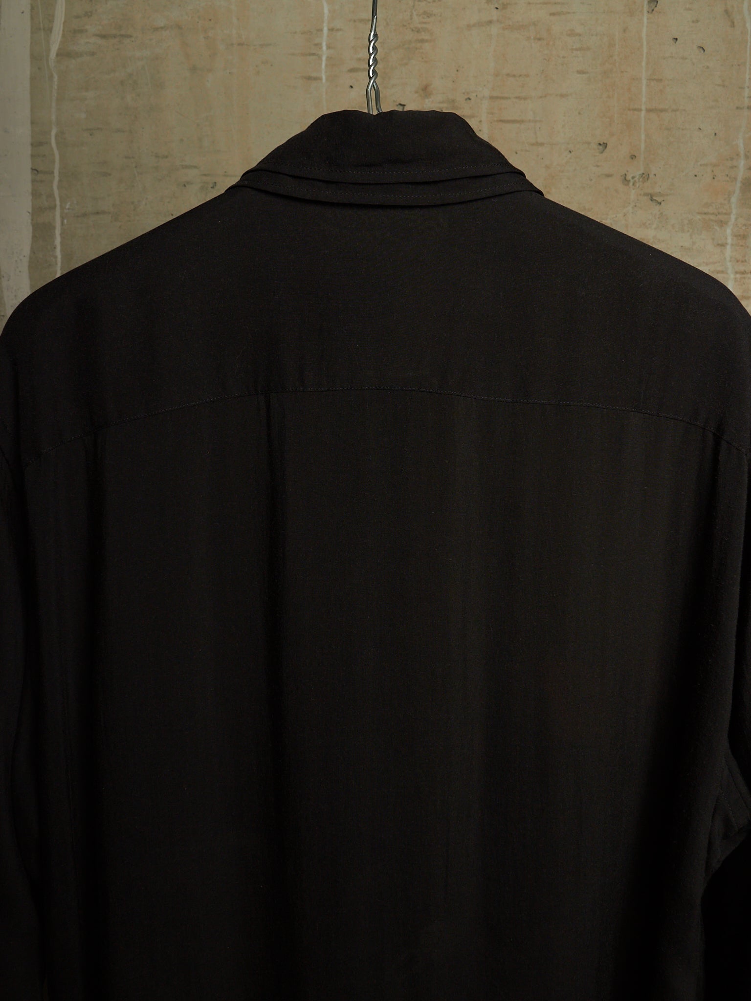 A black long-sleeve collared shirt hanging on a metal hanger against a textured beige wall, shown from the back.