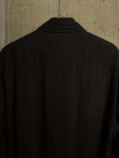 A black long-sleeve collared shirt hanging on a metal hanger against a textured beige wall, shown from the back.