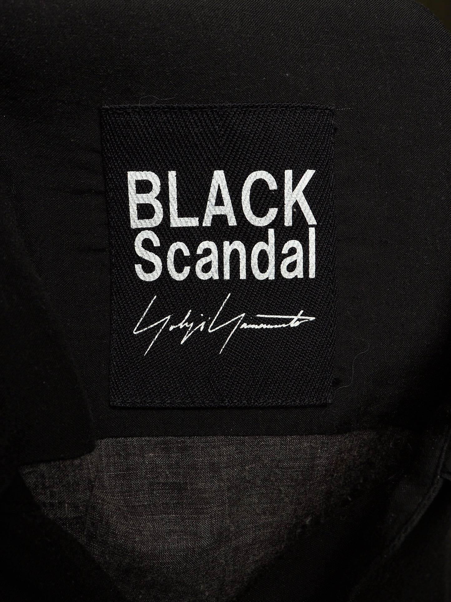Close-up of a black fabric patch with white embroidered text reading 'BLACK Scandal' and a white signature-style logo, attached to a black garment.
