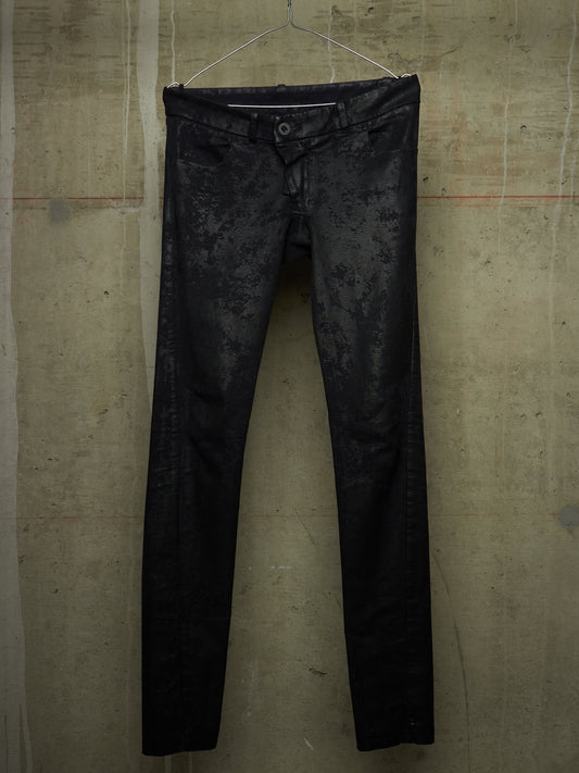 Black slim-fit jeans with a subtle tonal floral jacquard pattern, hanging on a wire hanger against a textured concrete wall.