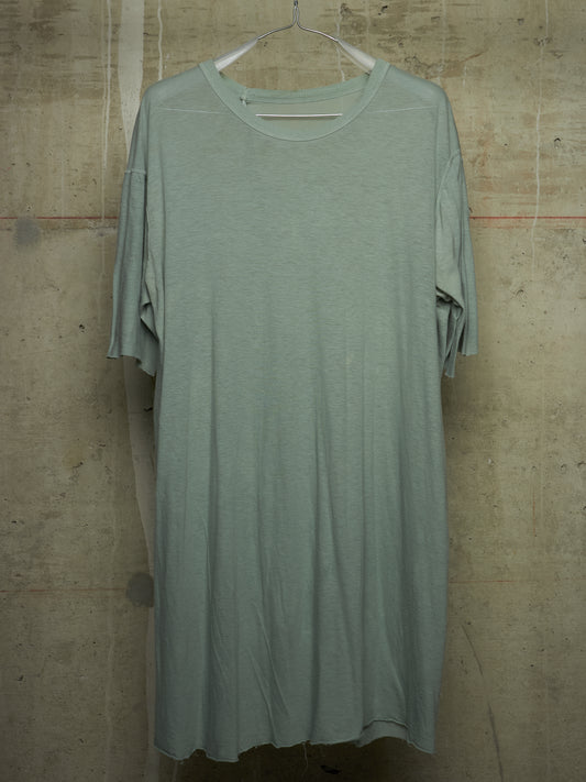 A loose, short-sleeve t-shirt dress in a muted sage green color, hanging on a white plastic hanger against a textured concrete wall.