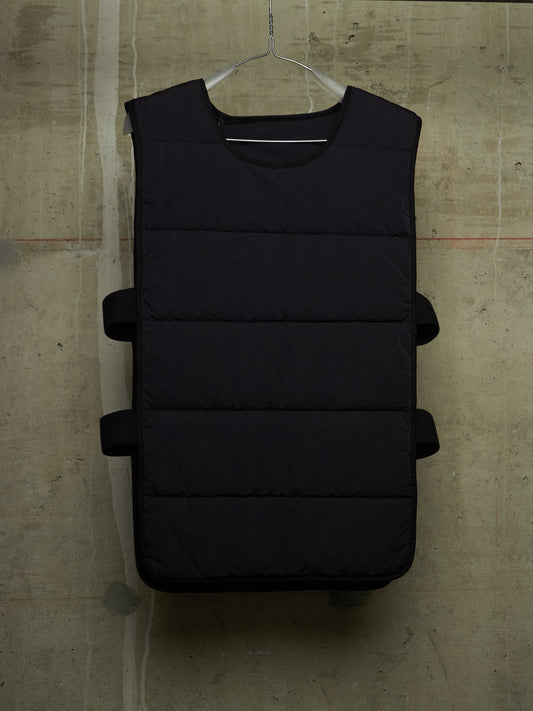 A black sleeveless puffer vest with horizontal quilted baffles and side straps, hanging on a metal hanger against a textured concrete wall.