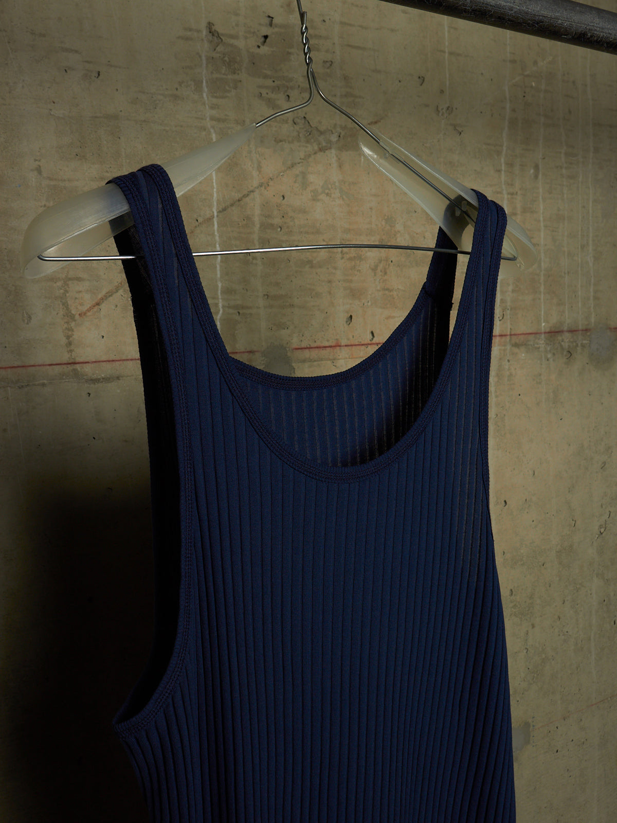 A dark navy blue ribbed tank top hanging on a clear plastic hanger against a textured concrete wall.