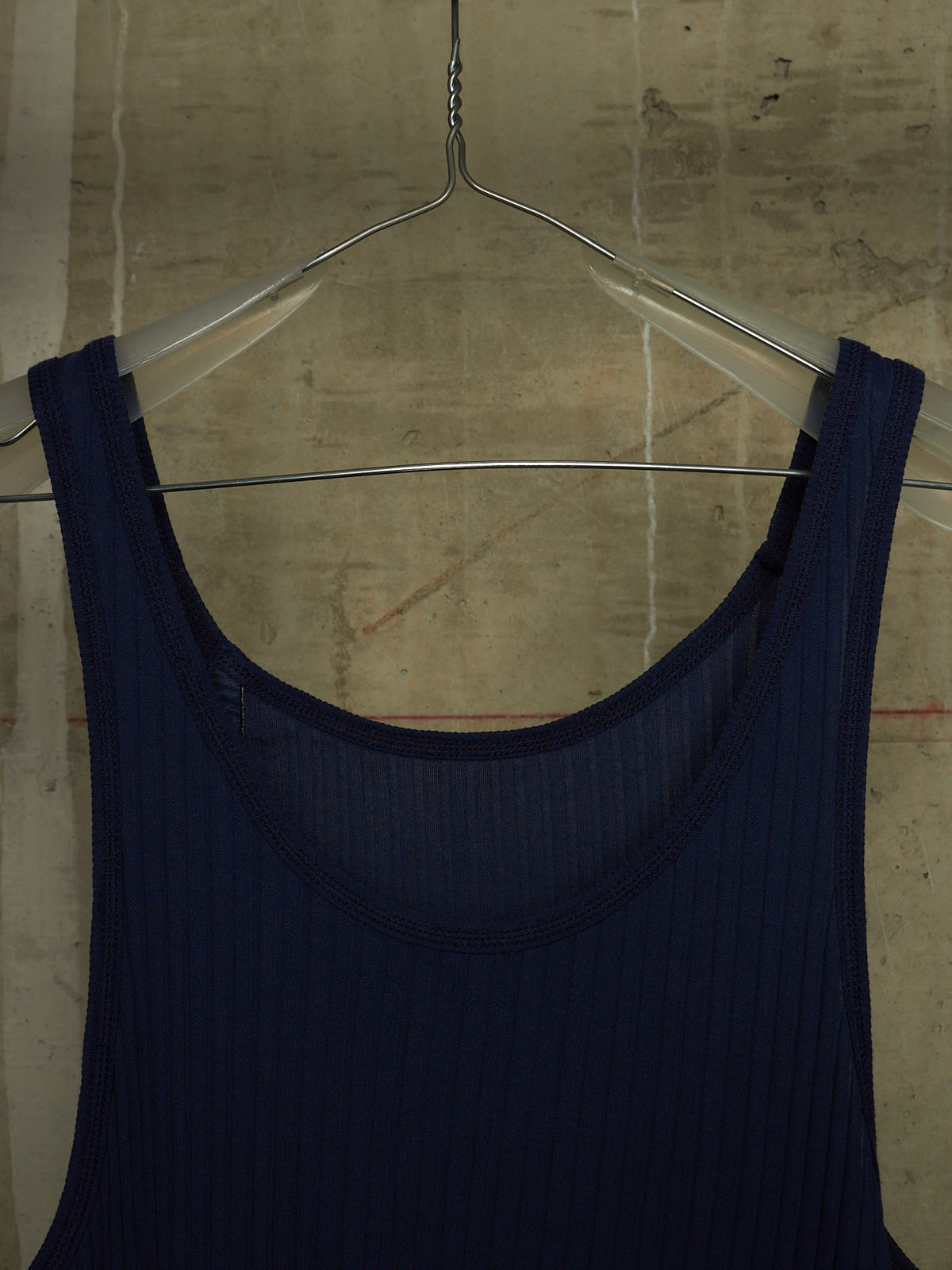 A dark navy blue ribbed tank top hanging on a clear plastic hanger against a textured concrete wall.