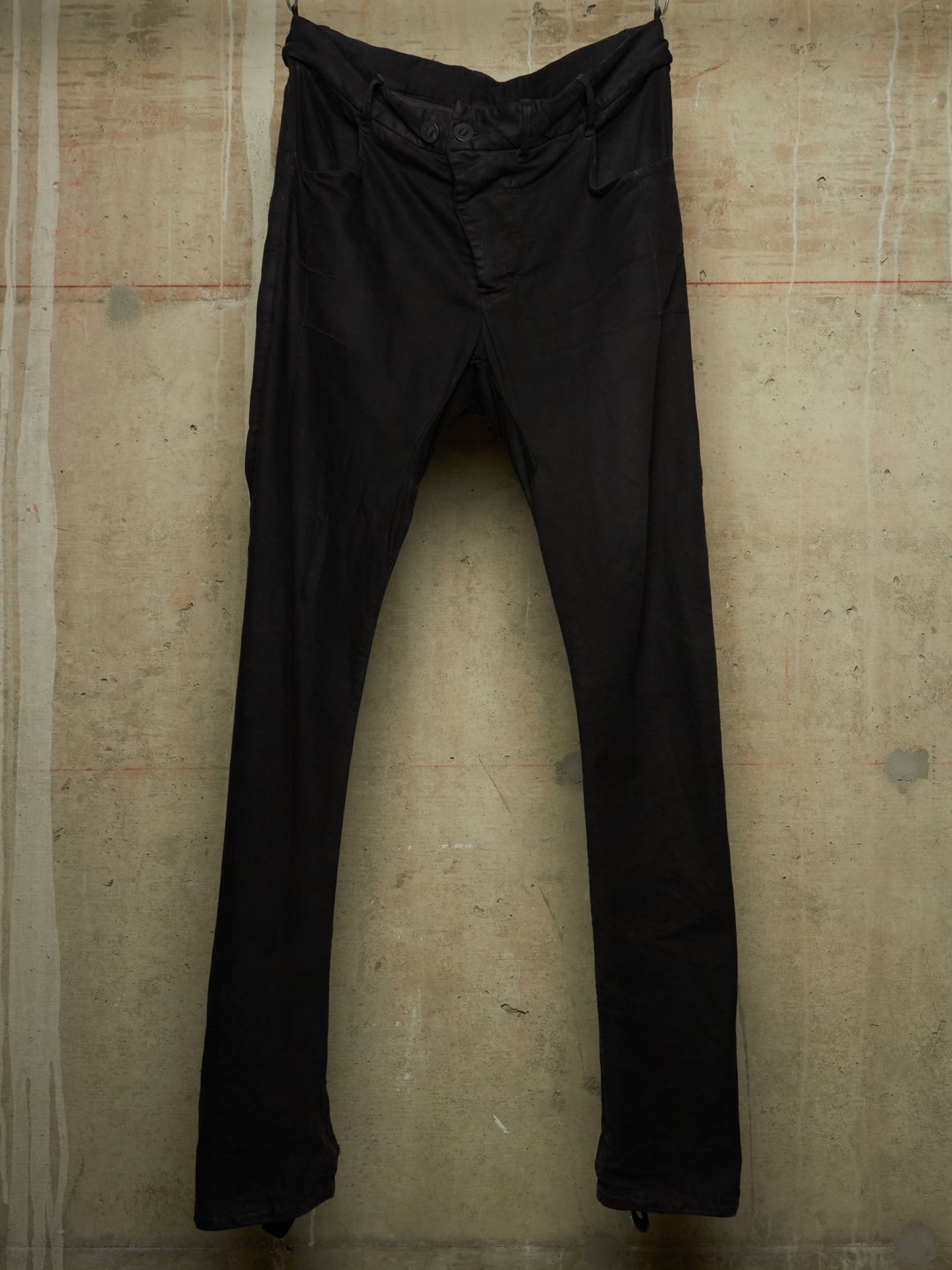 A pair of black slim-fit pants hanging against a textured concrete wall.