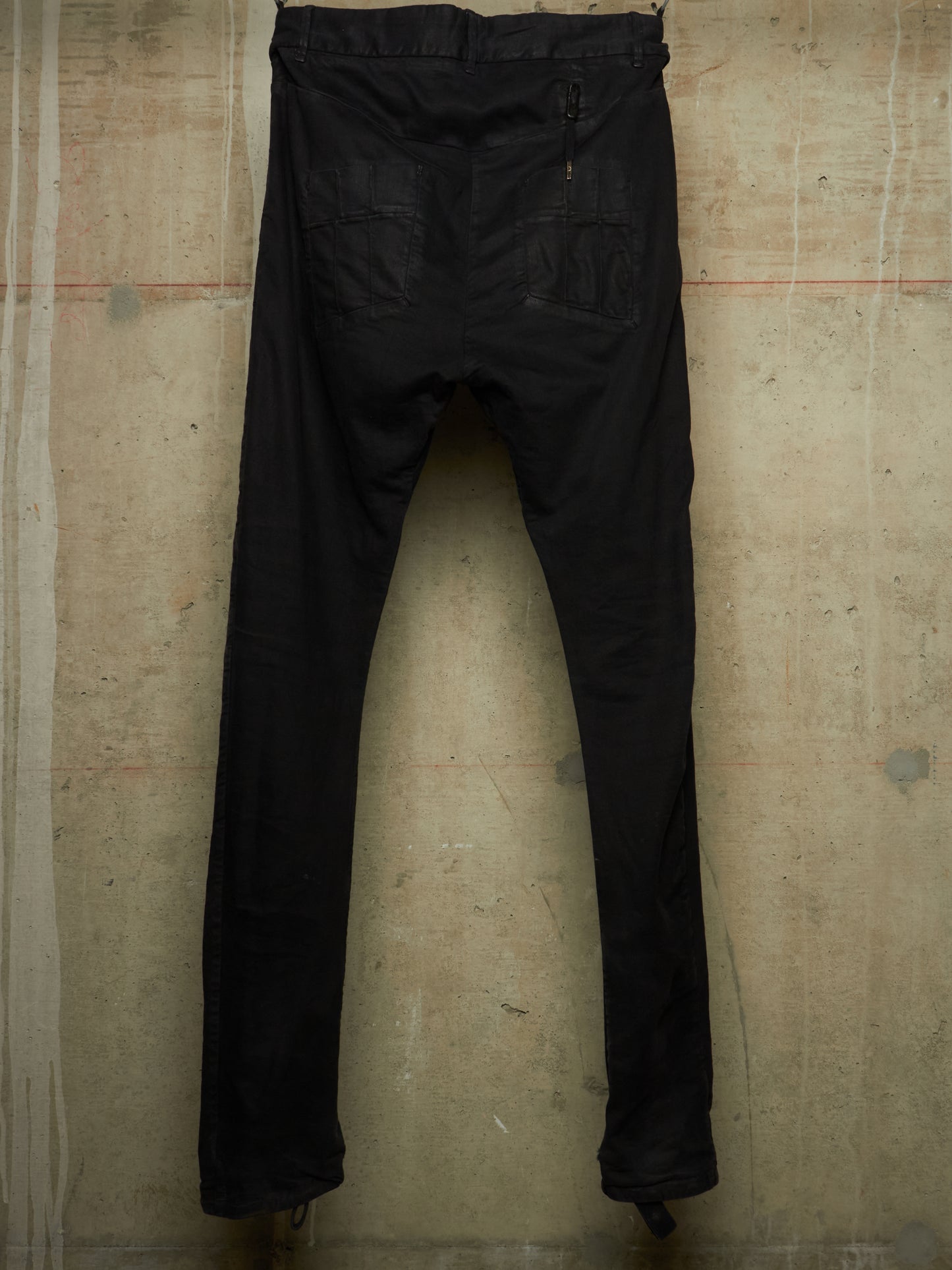Black slim-fit jeans shown from the back, hanging against a textured concrete wall.
