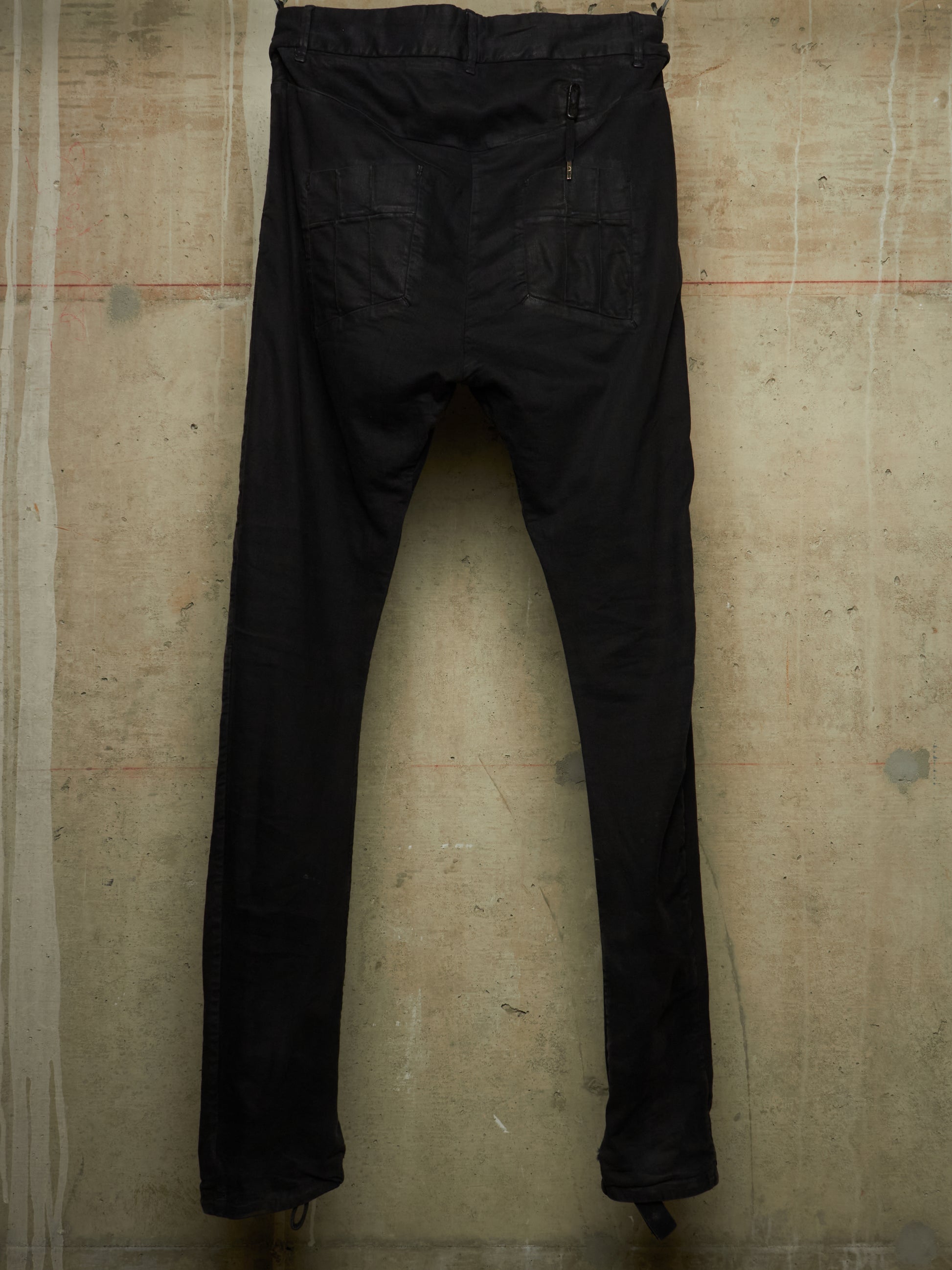 Black slim-fit jeans shown from the back, hanging against a textured concrete wall.