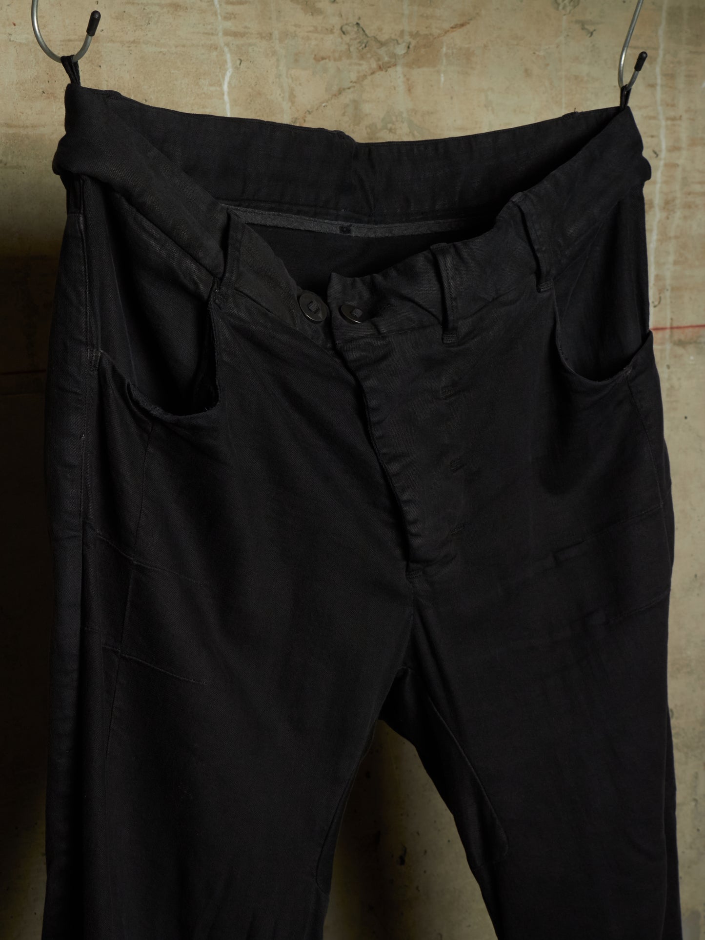 Black cotton trousers hanging from metal hooks against a textured concrete wall.