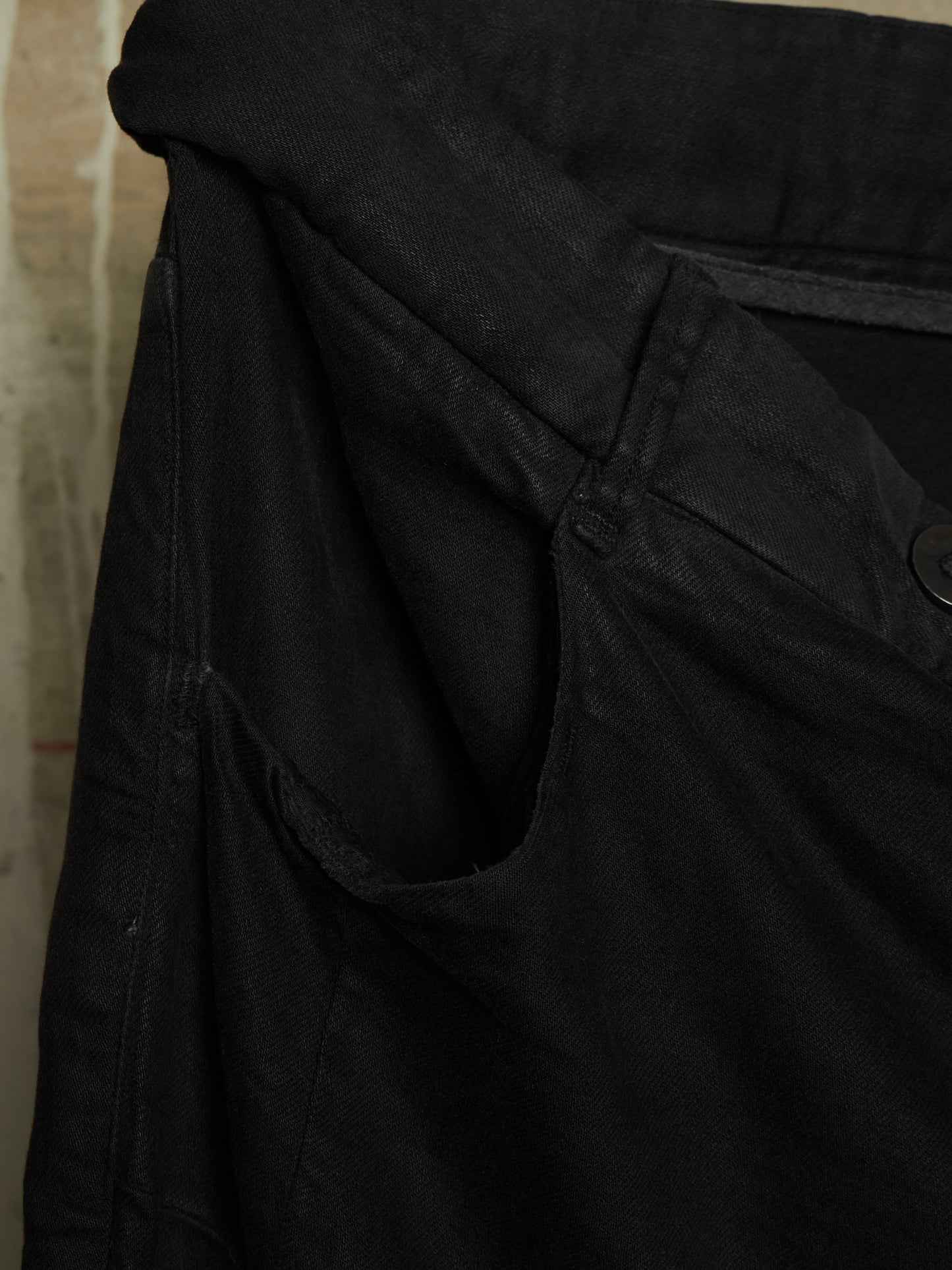 Close-up detail shot of a black denim garment, showing the waistband, pocket seam, and a dark button on a textured fabric background.