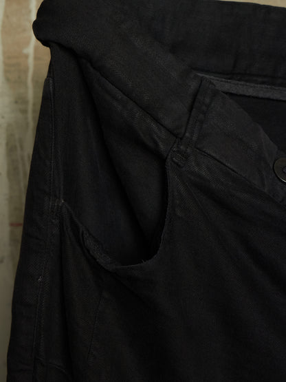 Close-up detail shot of a black denim garment, showing the waistband, pocket seam, and a dark button on a textured fabric background.
