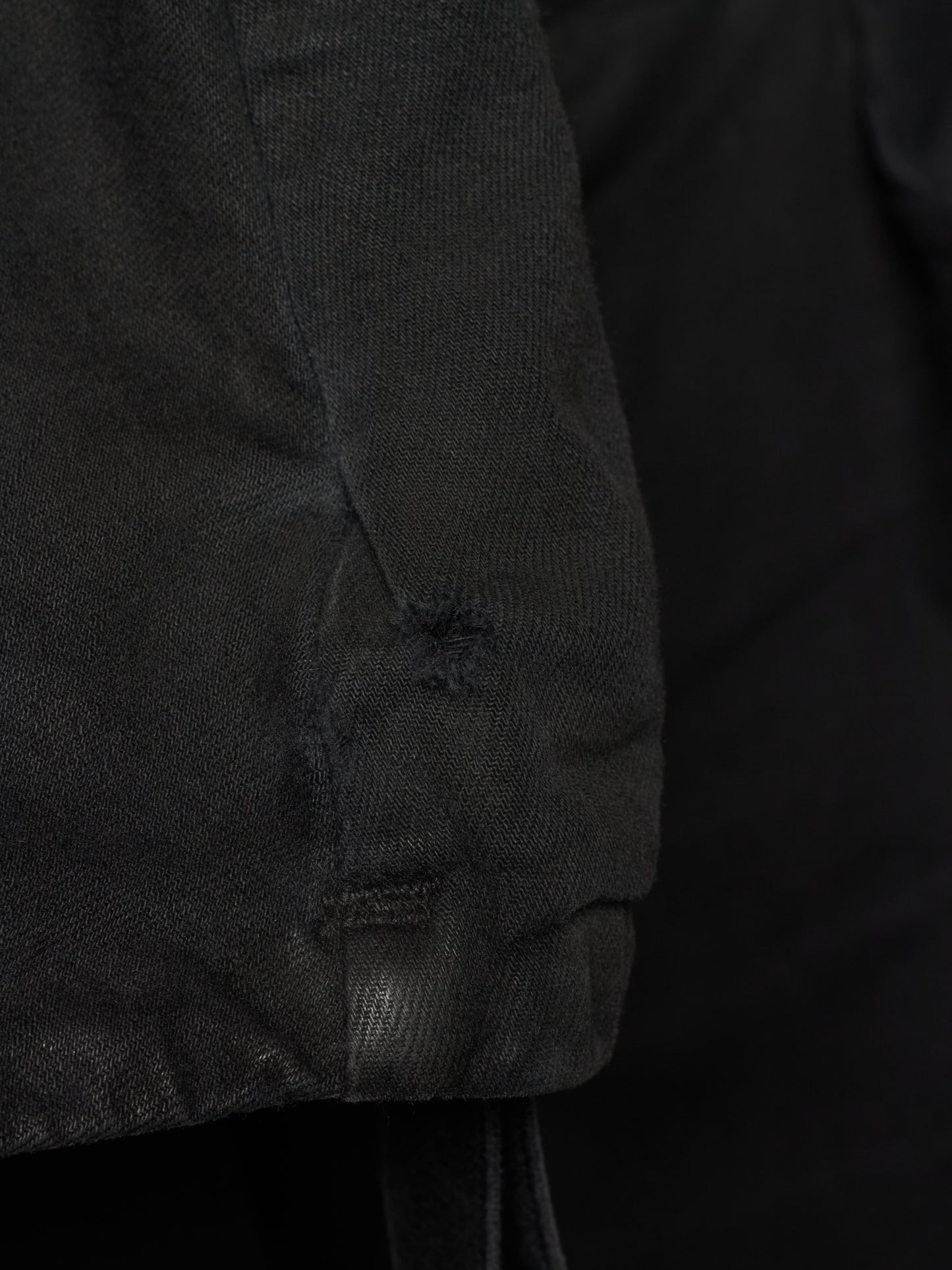 Close-up detail of a black textured garment, showing a sleeve or cuff with a small distressed hole and drawstring attachment against a dark background.