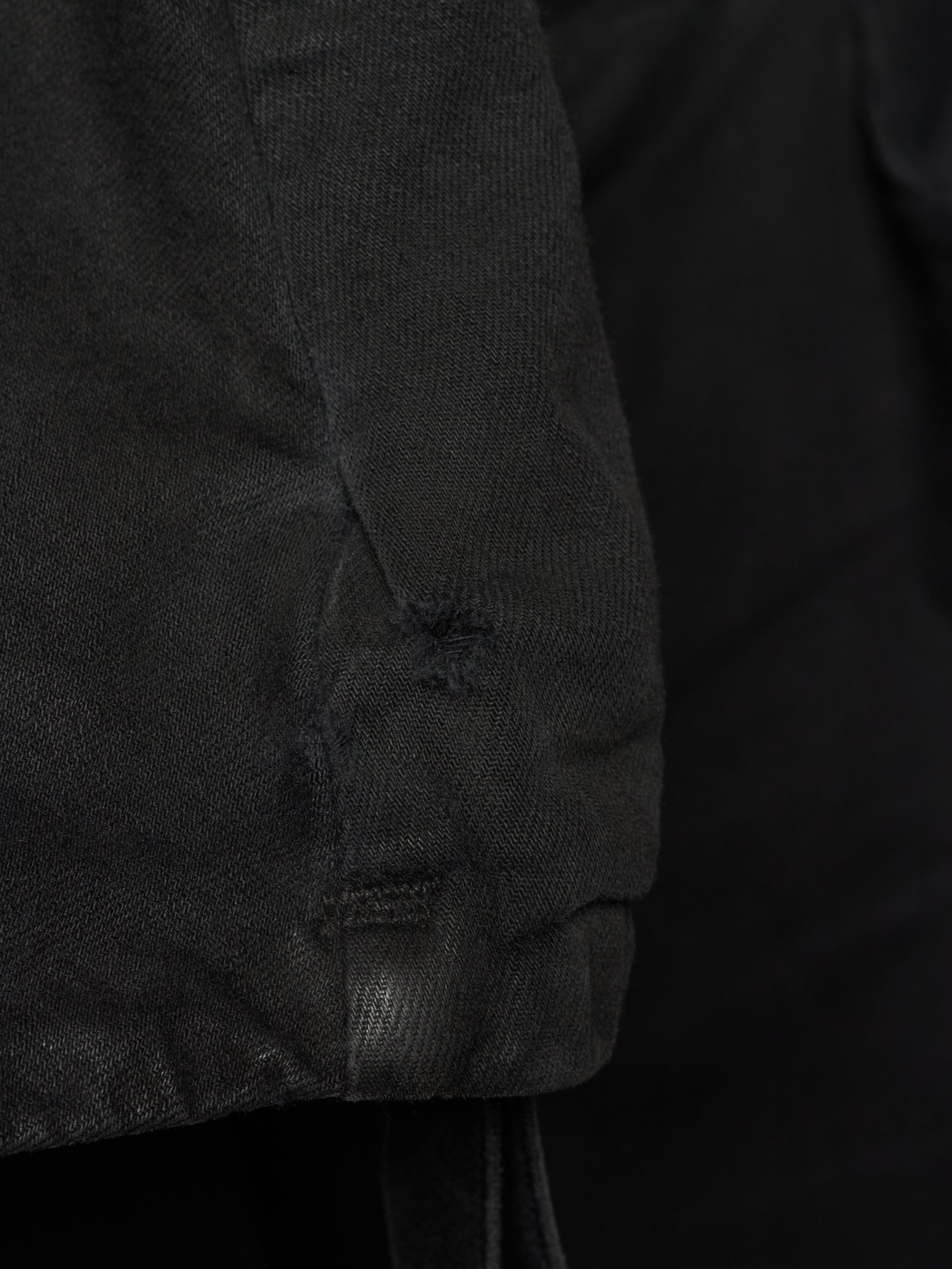 Close-up detail of a black textured garment, showing a sleeve or cuff with a small distressed hole and drawstring attachment against a dark background.