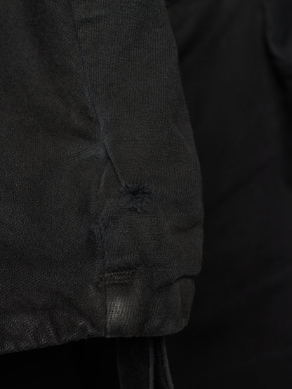 Close-up detail of a black textured garment, showing a sleeve or cuff with a small distressed hole and drawstring attachment against a dark background.