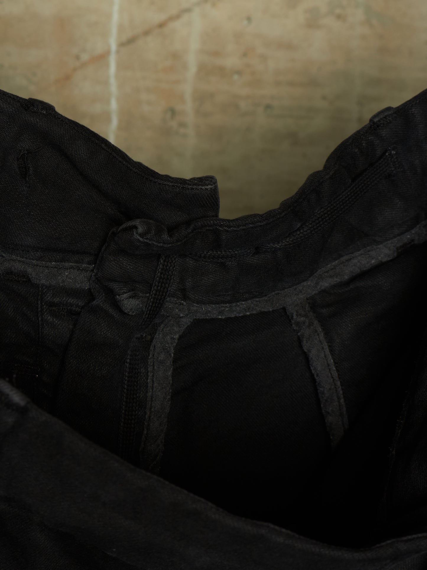 Close-up detail of a black garment, showing the inner waistband, zipper, and dark fabric texture against a warm beige background.
