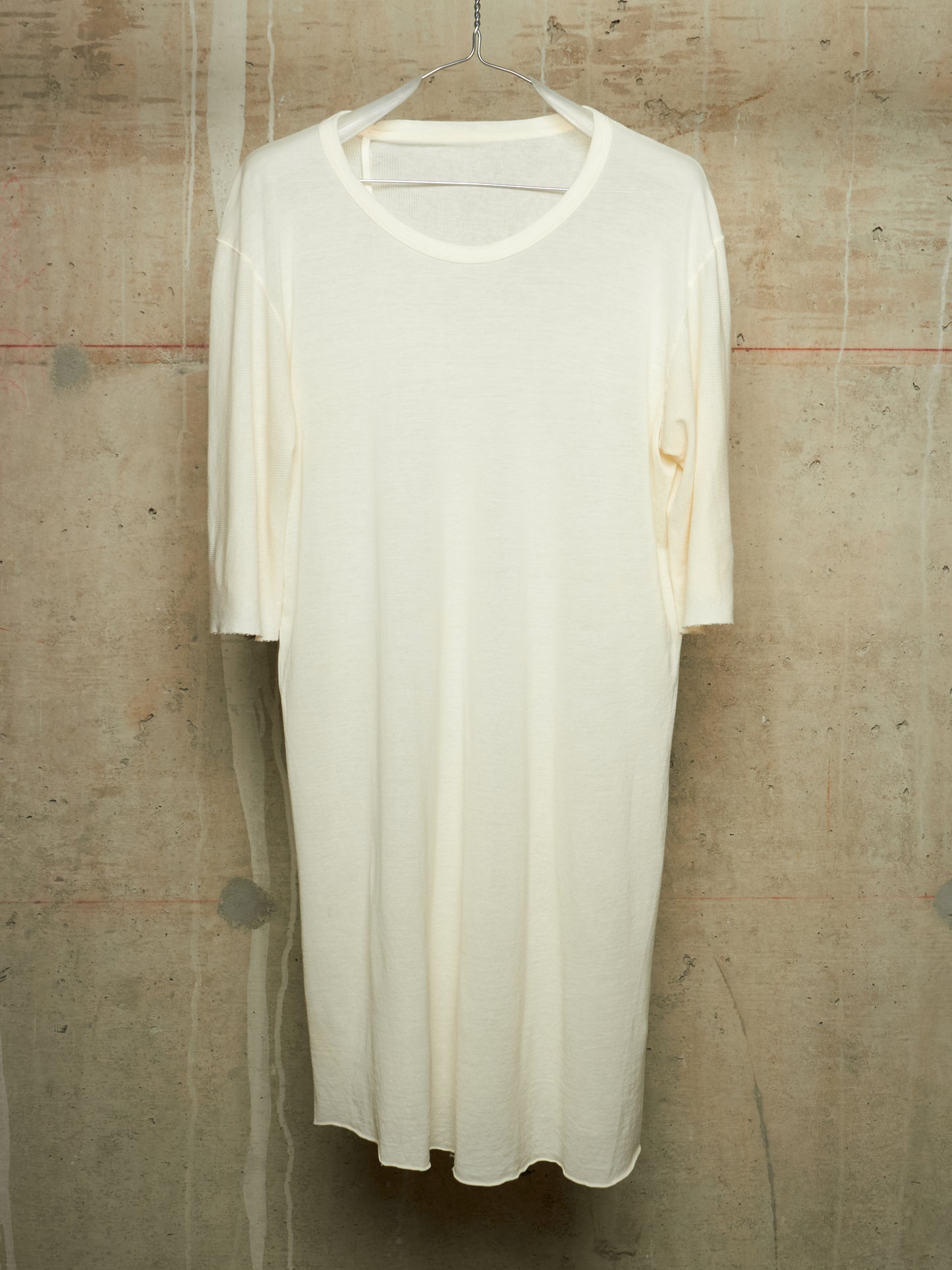 An off-white, semi-sheer short-sleeve tunic dress hanging on a metal hanger against a textured concrete wall.