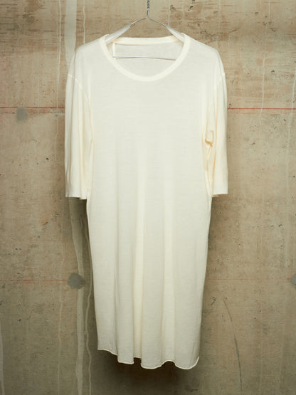 An off-white, semi-sheer short-sleeve tunic dress hanging on a metal hanger against a textured concrete wall.