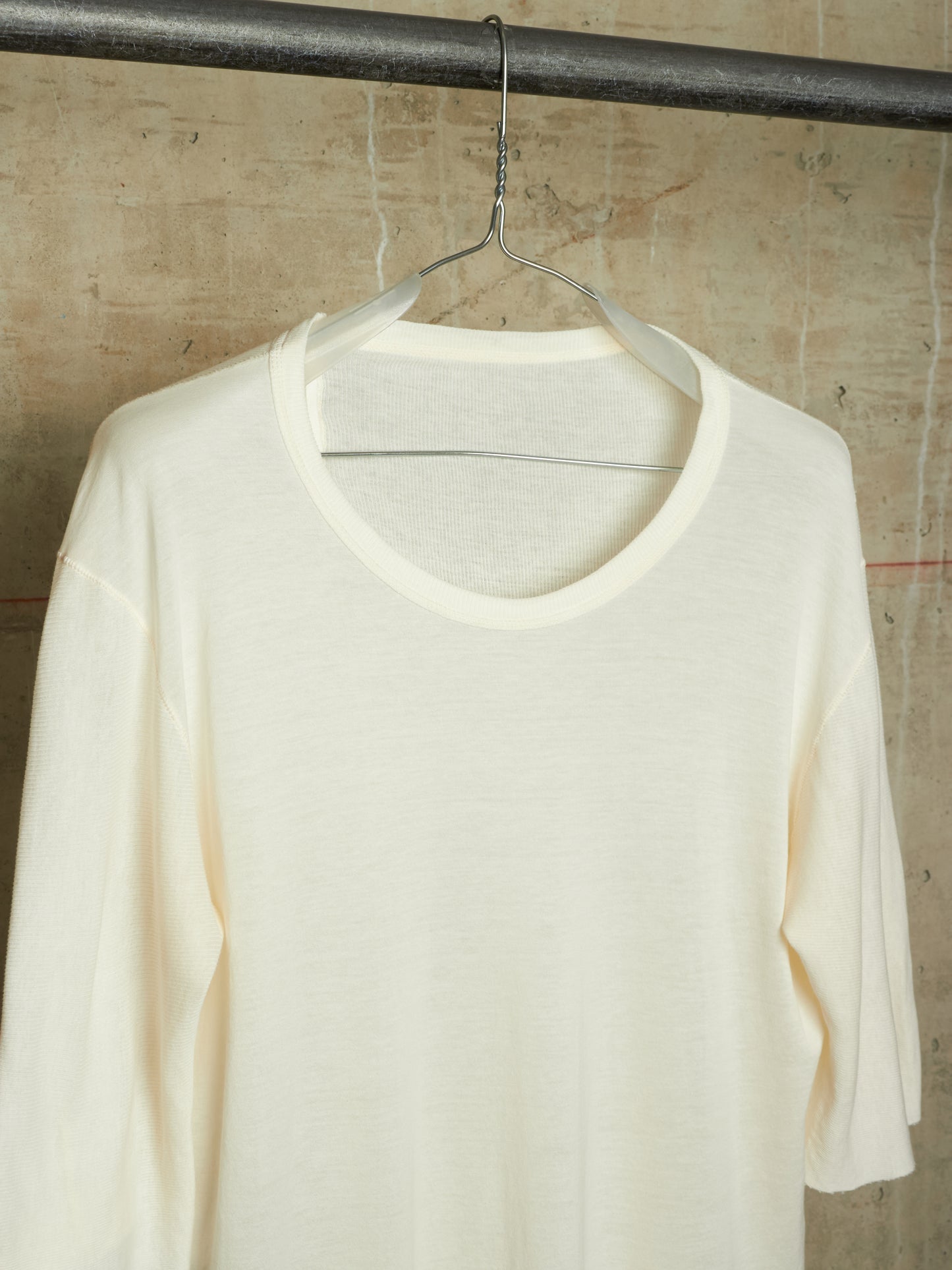 An off-white, cream-colored long-sleeve knit top hanging on a clear plastic hanger from a dark metal rod, set against a textured concrete wall.