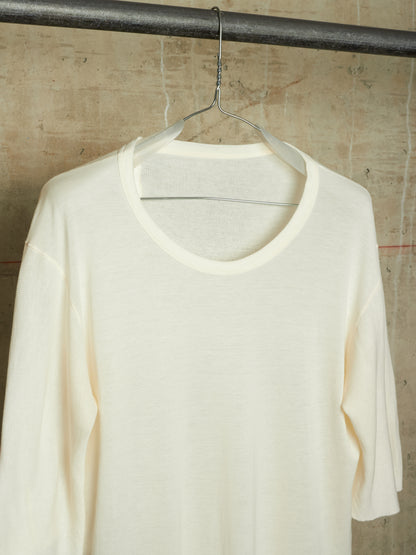 An off-white, cream-colored long-sleeve knit top hanging on a clear plastic hanger from a dark metal rod, set against a textured concrete wall.