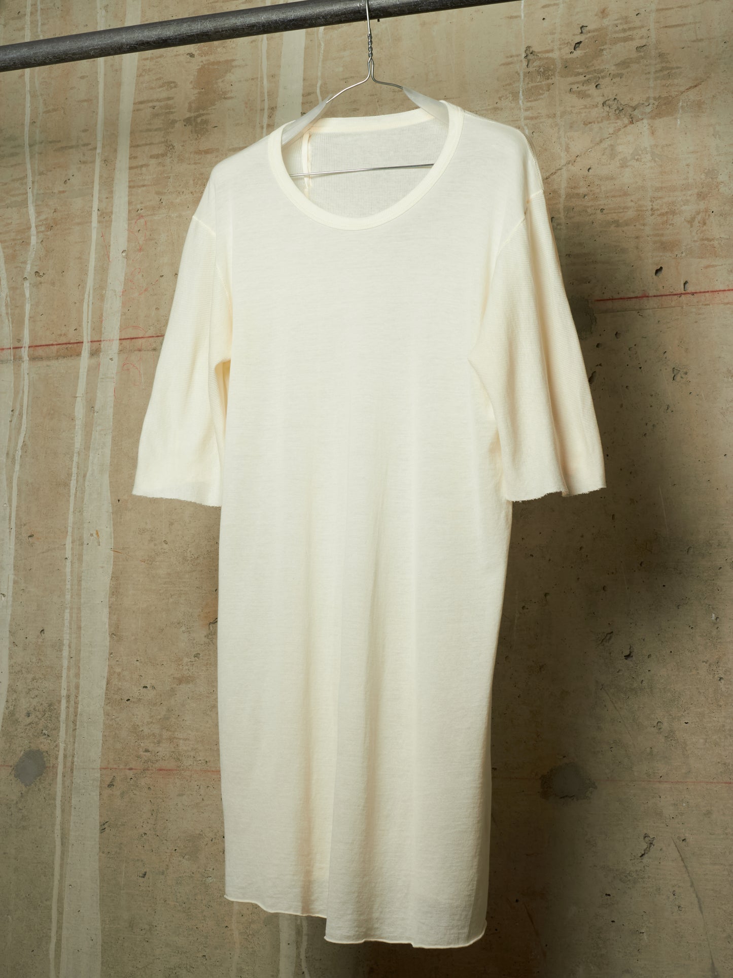An off-white, cream-colored short-sleeve knit dress or tunic hanging on a wire hanger from a dark metal rod, set against a textured concrete wall.