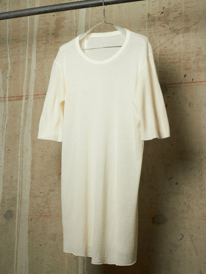 An off-white, cream-colored short-sleeve knit dress or tunic hanging on a wire hanger from a dark metal rod, set against a textured concrete wall.