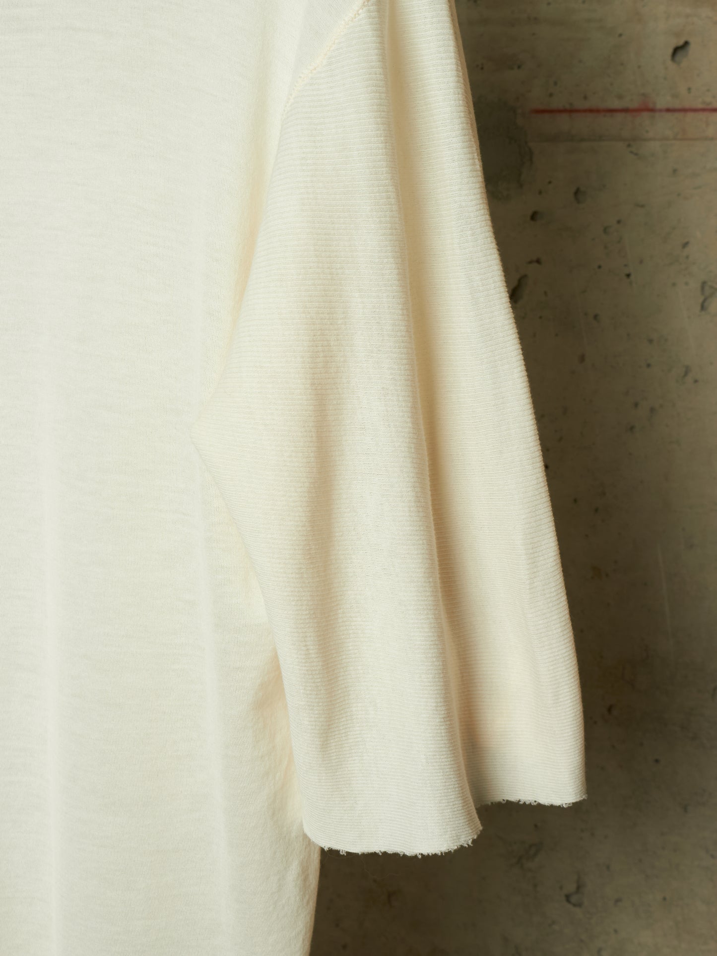 A close-up of a cream-colored knit garment, showing the fine ribbed texture of the sleeve and body, set against a textured concrete background.