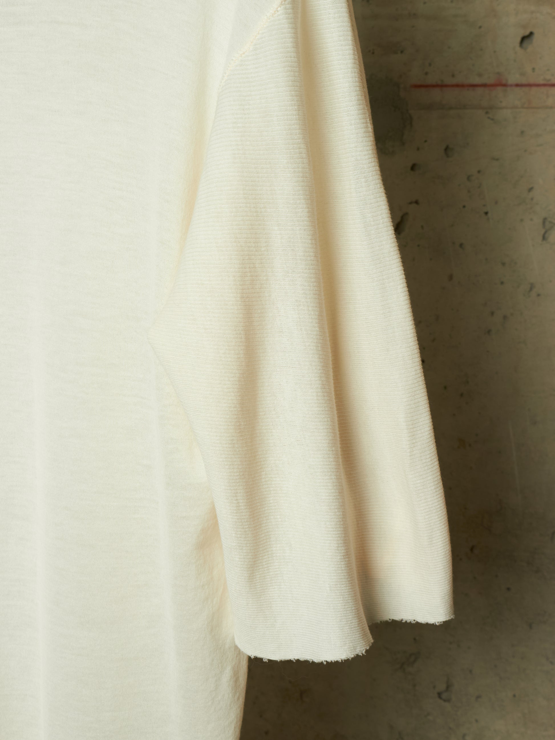 A close-up of a cream-colored knit garment, showing the fine ribbed texture of the sleeve and body, set against a textured concrete background.