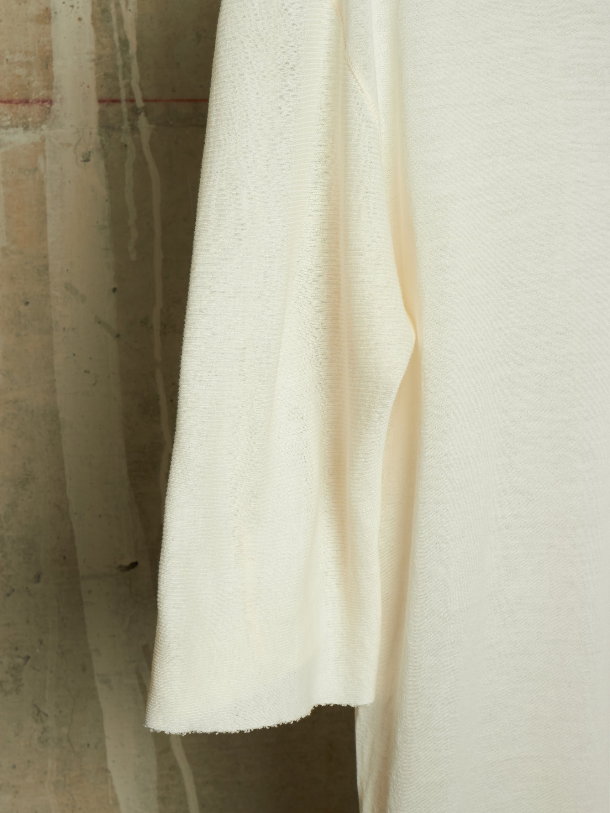 A close-up of a cream-colored knit fabric garment, showing the texture and drape of the material against a neutral concrete background.