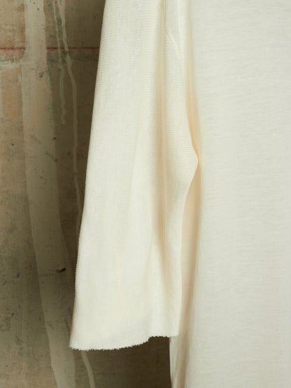 A close-up of a cream-colored knit fabric garment, showing the texture and drape of the material against a neutral concrete background.