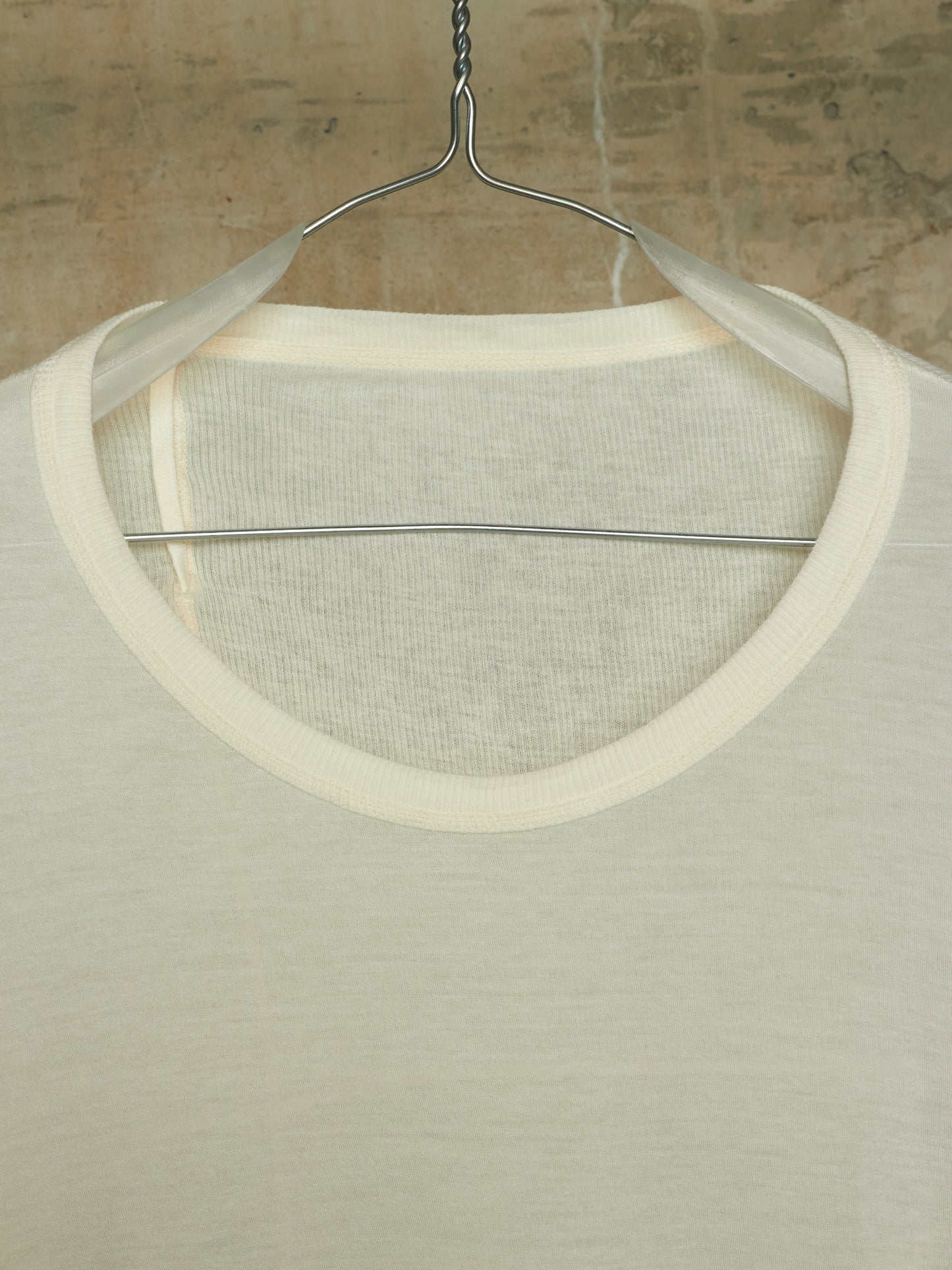 A close-up of a cream-colored knit top hanging on a metal hanger against a textured, warm-toned wall.