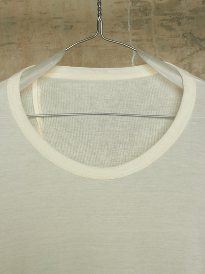 A close-up of a cream-colored knit top hanging on a metal hanger against a textured, warm-toned wall.