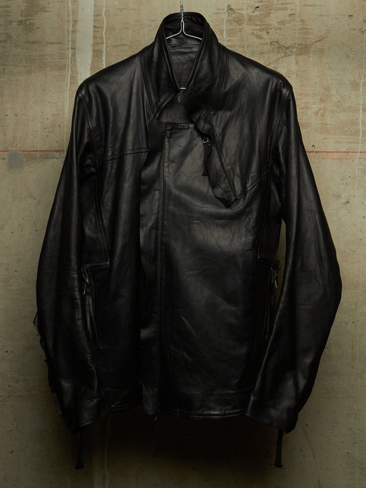 A black leather jacket with a high collar and asymmetrical zipper closure, hanging on a wire hanger against a textured concrete wall.