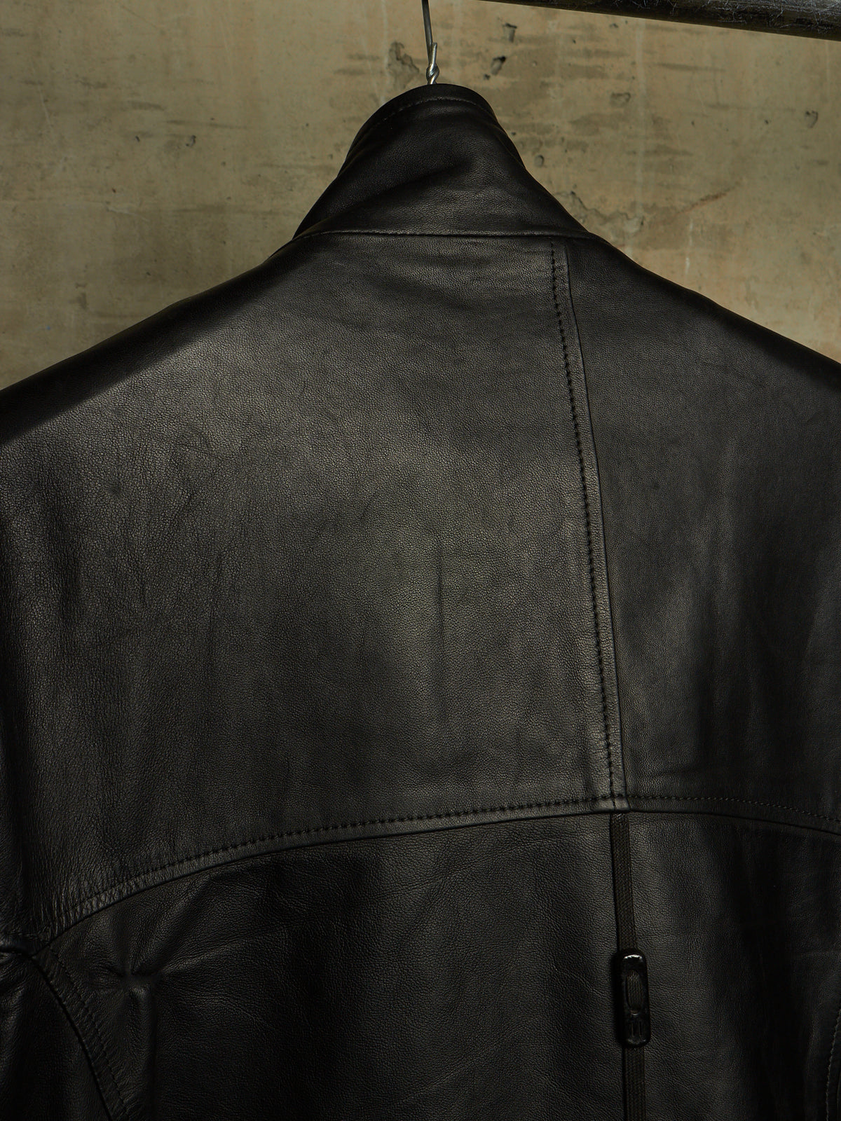 Close-up of the back of a black leather jacket hanging against a textured concrete wall.
