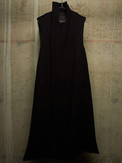 A sleeveless black wrap-style maxi dress with a high mock neck, hanging on a black hanger against a textured concrete wall.