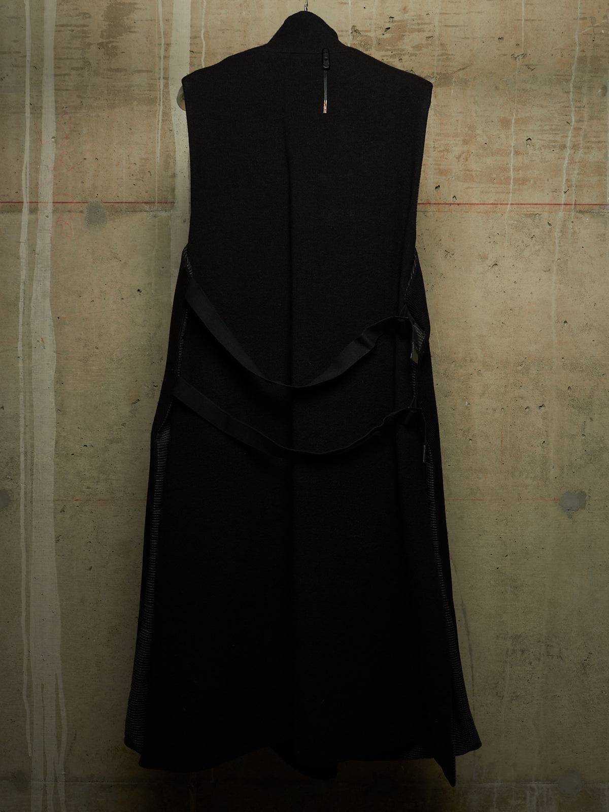 A sleeveless black dress with a high collar and layered ruffle details, shown hanging against a textured concrete wall.