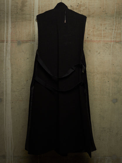 A sleeveless black dress with a high collar and layered ruffle details, shown hanging against a textured concrete wall.