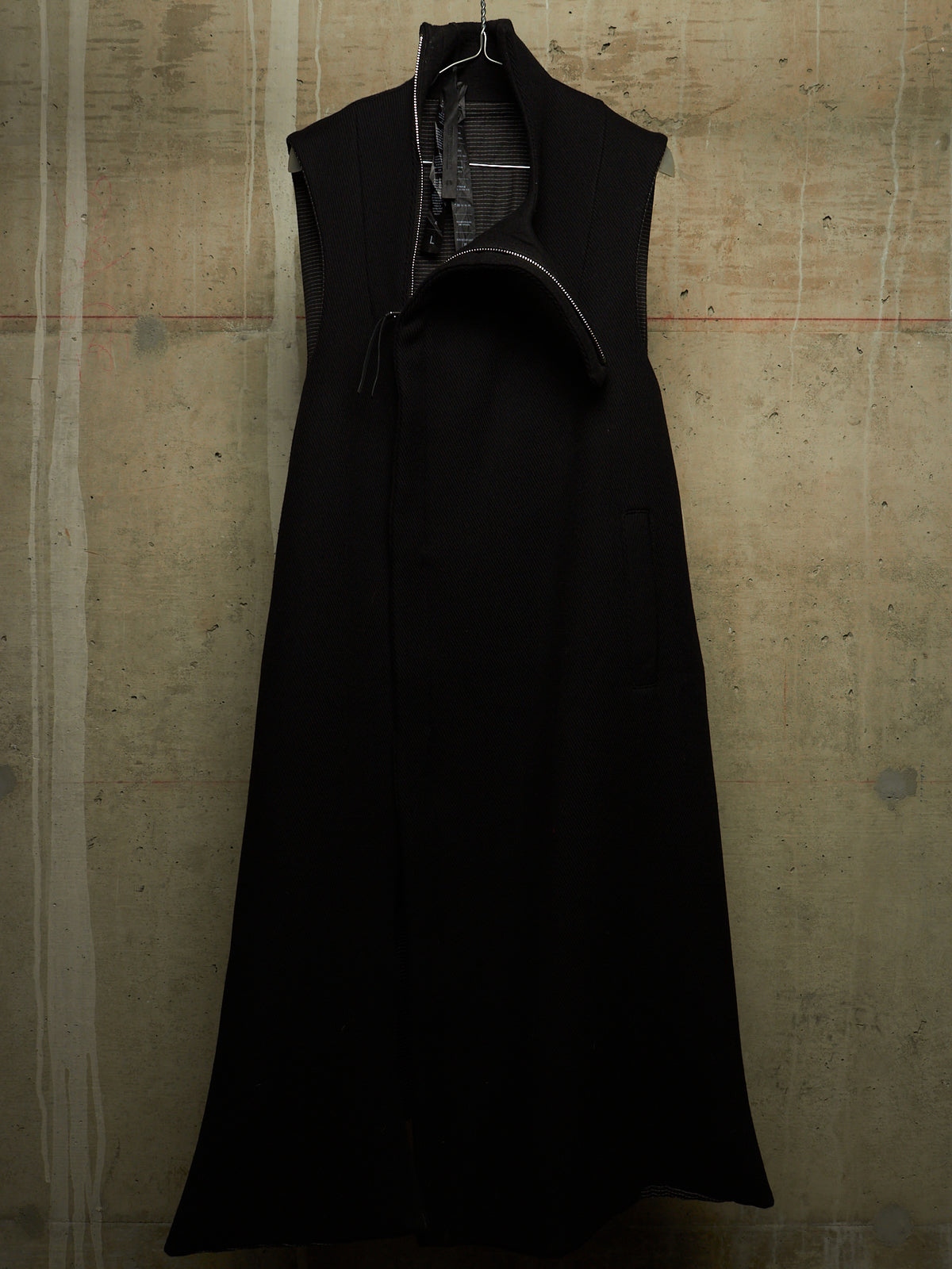 A black sleeveless long coat with a high collar and asymmetrical front opening, hanging against a textured concrete wall.